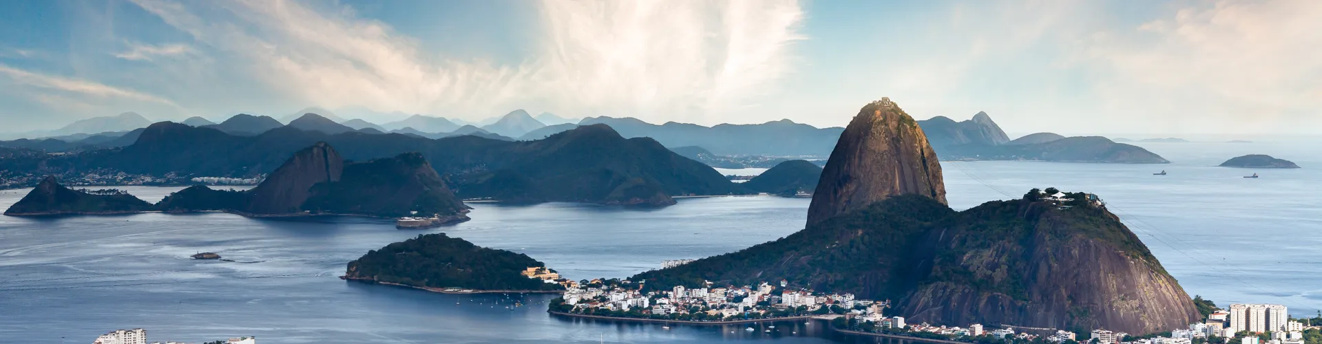 Panoramic bay view with hills, mountains, a large rock formation, and coastal buildings.