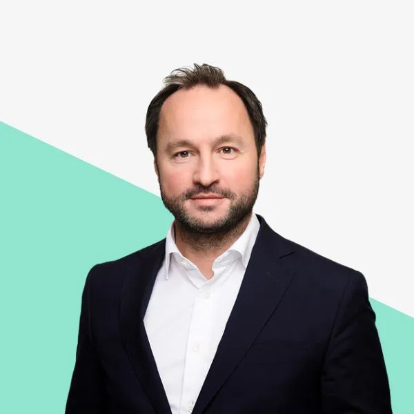 Man with dark hair and beard in a navy suit against a plain background with a green triangle.