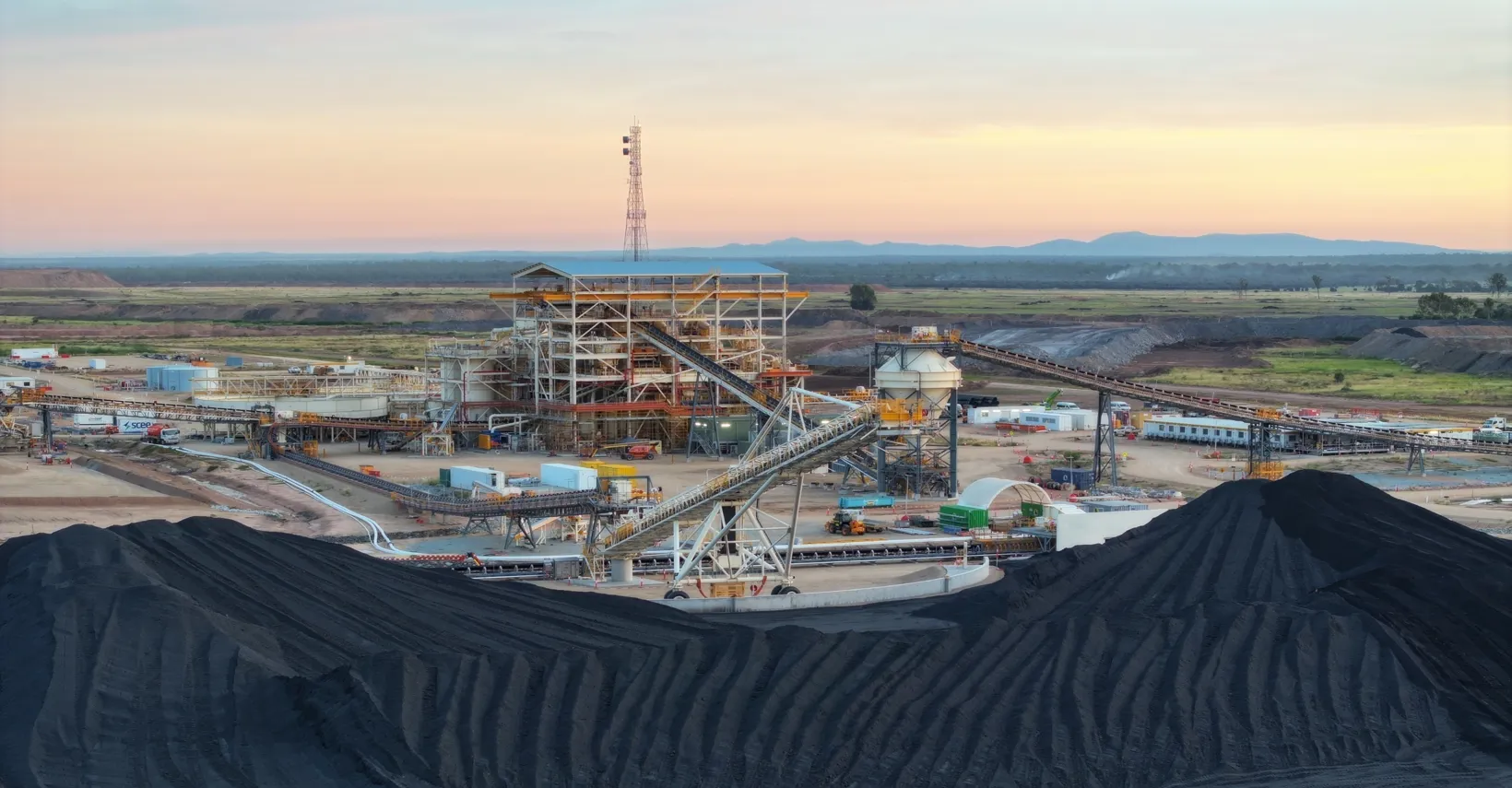 Coal processing facility with coal piles, conveyor belts, and machinery at sunrise.