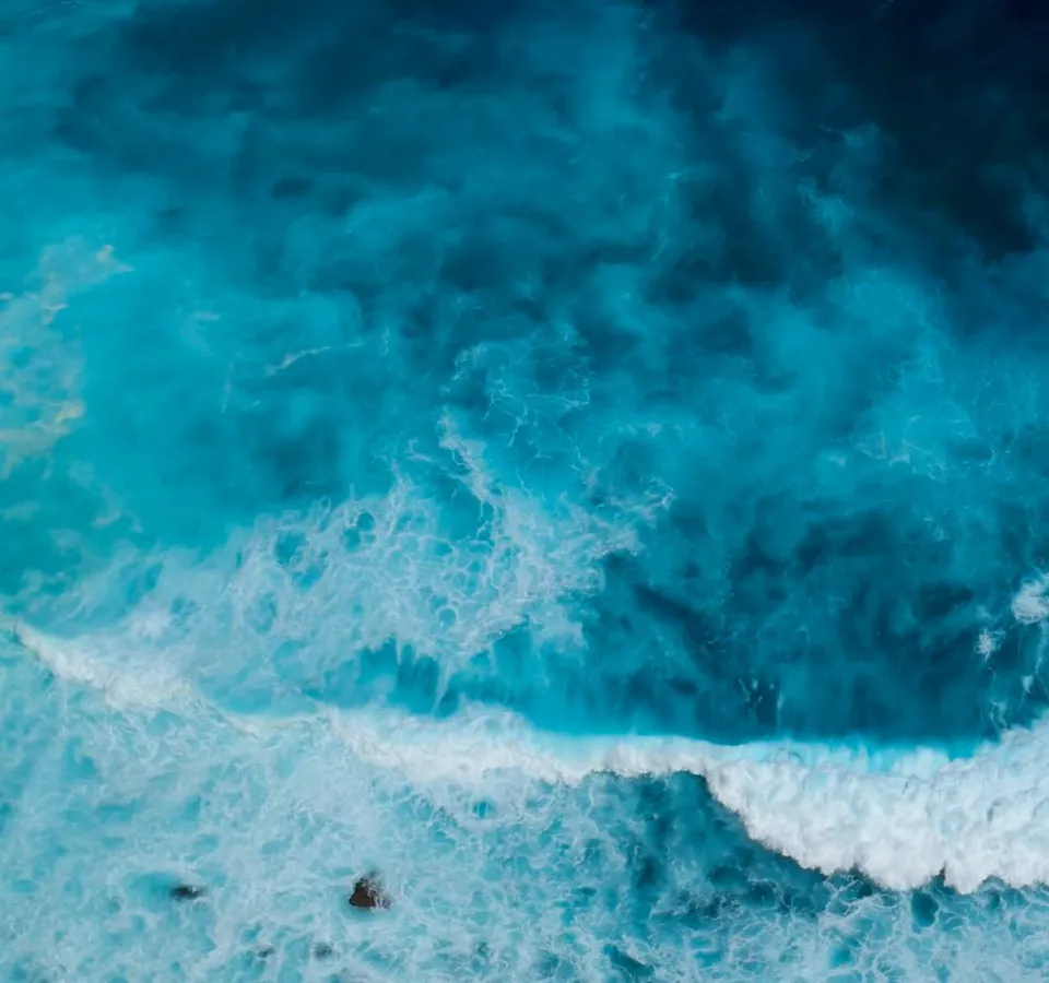 Aerial view of turquoise ocean with white waves crashing near rocky shore.