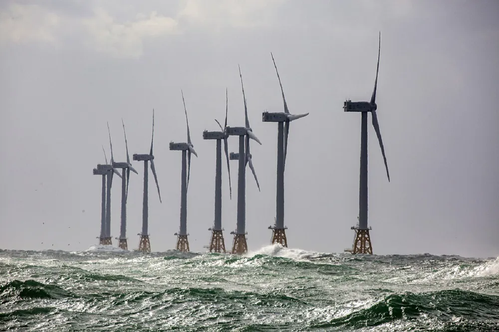 Row of large offshore wind turbines under gray skies with choppy waves.