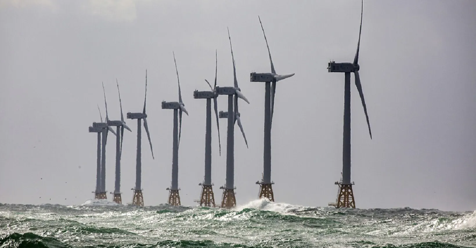 Row of large offshore wind turbines under gray skies with choppy waves.