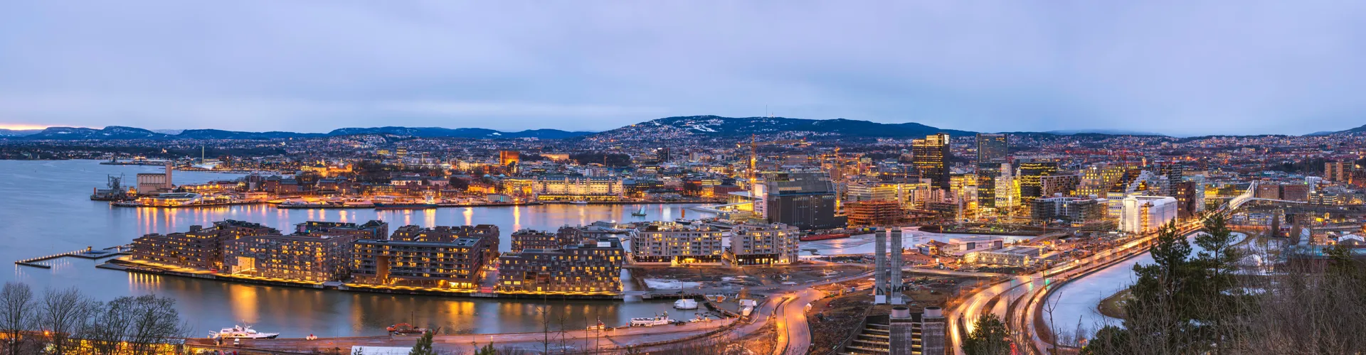Panoramic twilight view of a city with illuminated buildings, water, and boats.
